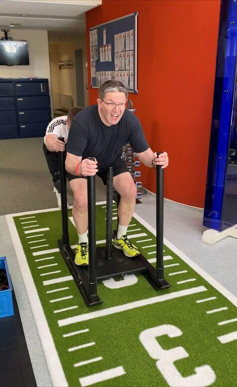 Mann schiebt einen Schlitten auf einer grünen Sportmatte, während ein weiterer Mensch ihn von hinten unterstützt.