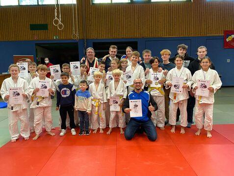 Gruppe von Jugendlichen in Judoanzügen hält Urkunden, umgeben von Trainern in einer Judohalle mit roten Matten.