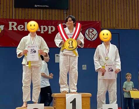 Drei Jugendliche in Judoanzügen stehen auf dem Siegertreppchen, der Mittelsteher hält eine Goldmedaille und Urkunde.