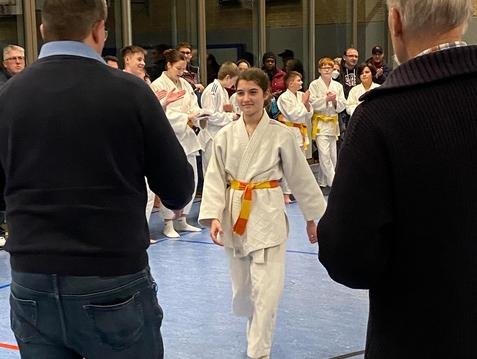 Junge Kämpferin in Karate-Uniform mit orangefarbener Gürtelschärfe geht auf eine Gruppe applaudierender Zuschauer zu.