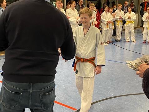 Junge im Karateanzug mit orangem Gürtel schüttelt einem Erwachsenen die Hand, während Zuschauer applaudieren.