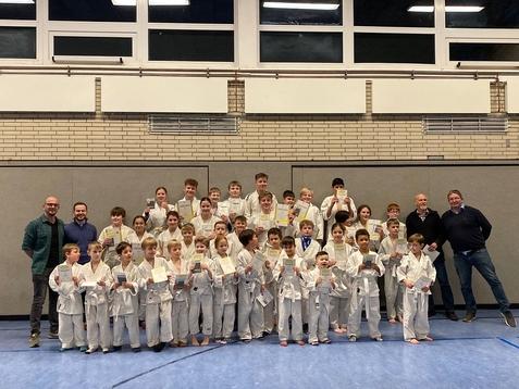 Gruppenfoto von Kindern in weißen Judogi mit Urkunden und begeisterten Trainern in einer Sporthalle.