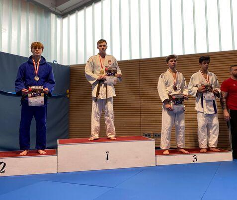 Vier Judokas stehen auf dem Podest, halten Urkunden und Medaillen, auf einer blauen Matte in einer Sporthalle.