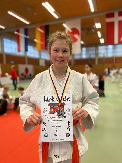 Mädchen in Judogi hält stolz eine Urkunde mit Medaille in einem Sportzentrum, im Hintergrund Flaggen und andere Teilnehmer.