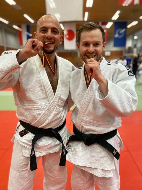 Zwei Kämpfer in Judoanzügen halten Siegermedaillen vor dem Gesicht und lächeln auf einer roten Matte.
