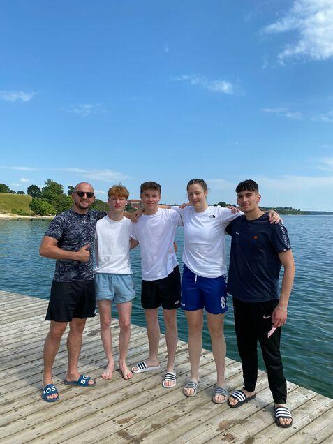 Fünf junge Menschen in sportlicher Kleidung stehen lächelnd auf einem Holzsteg am Wasser unter blauem Himmel.