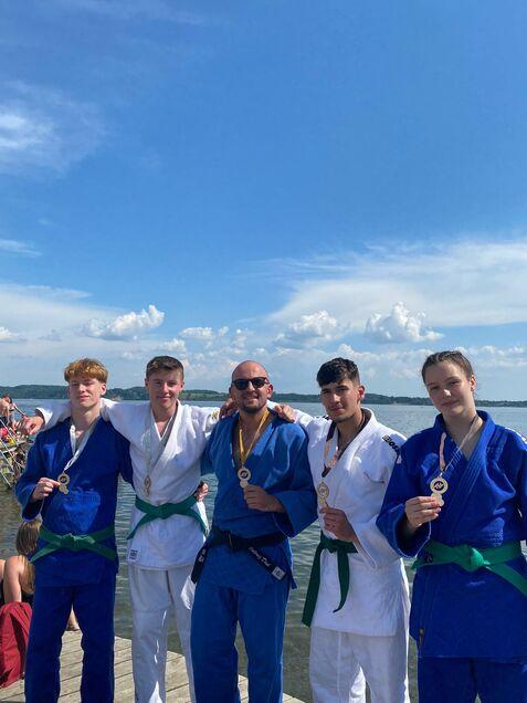 Fünf Judoka in Anzügen mit Medaillen posieren am Ufer eines Gewässers unter blauem Himmel.
