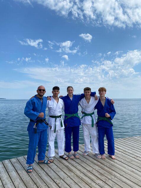 Fünf Personen in BJJ-Anzügen stehen auf einem Holzsteg am Wasser, lächeln in die Kamera unter blauem Himmel.