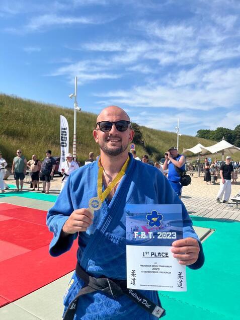 Mann in blauer Judo-Gi hält Medaille und Urkunde für den 1. Platz beim Federica Beach Turnier 2023 in der Hand.