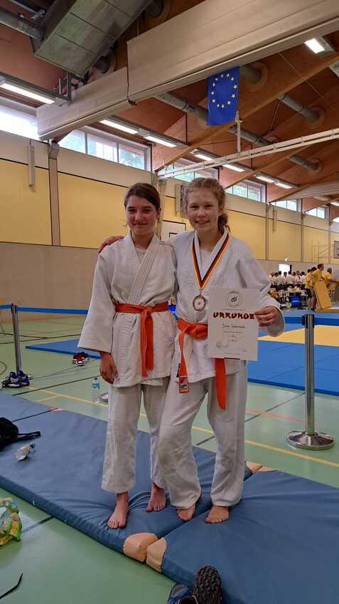Zwei Judo-Kämpferinnen in weißen Anzügen mit orangen Gürteln stehen auf einer Matte und halten eine Urkunde.