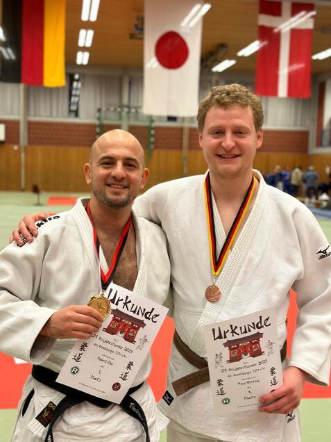 Zwei Judoka in weißen Anzügen halten Urkunden und Medaillen vor einer Sporthalle mit Flaggen im Hintergrund.