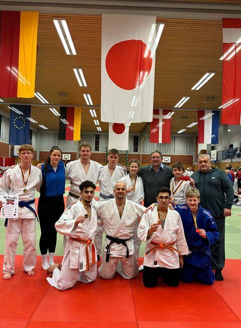 Gruppe von Judoka mit Medaillen in der Sporthalle, umgeben von Flaggen und Matten. Urkunden in Händen eines Teilnehmers.
