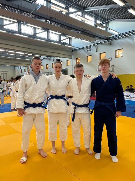 Vier Judoka in Trainingsanzügen mit blauen Gürteln posieren auf einer Judomatte in einer Halle.