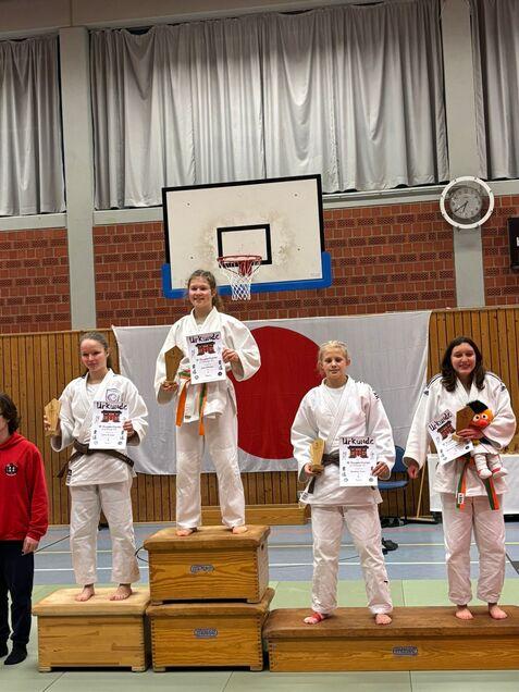 Vier Judokas auf dem Siegertreppchen halten Urkunden und Pokale in einer Sporthalle mit einem Basketballkorb im Hintergrund.