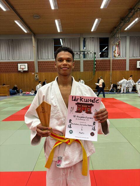 Junge im Judoanzug hält eine Urkunde und einen Pokal, neben ihm Judo-Turnier-Hintergrund mit Matten und Teilnehmern.