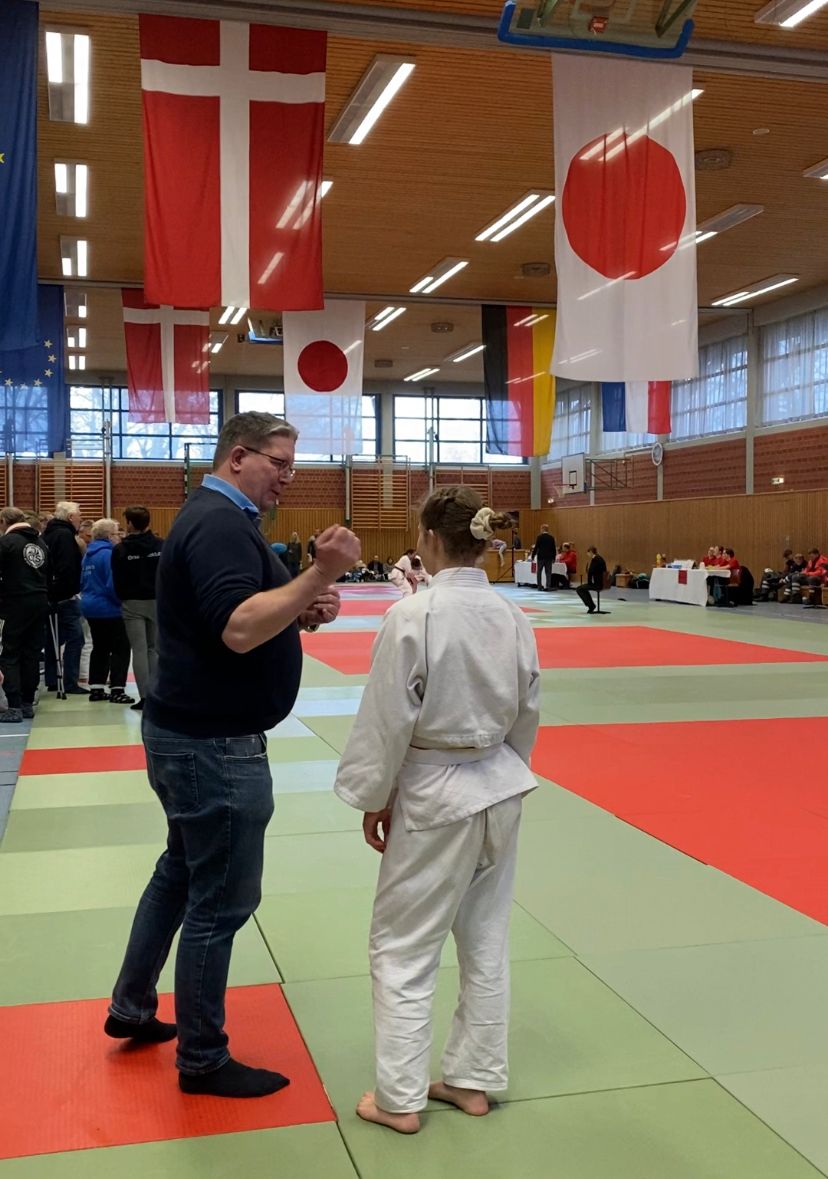 Ein Trainer spricht mit einem jungen Judoka in einem Sportzentrum, umgeben von Flaggen und anderen Wettkämpfern.