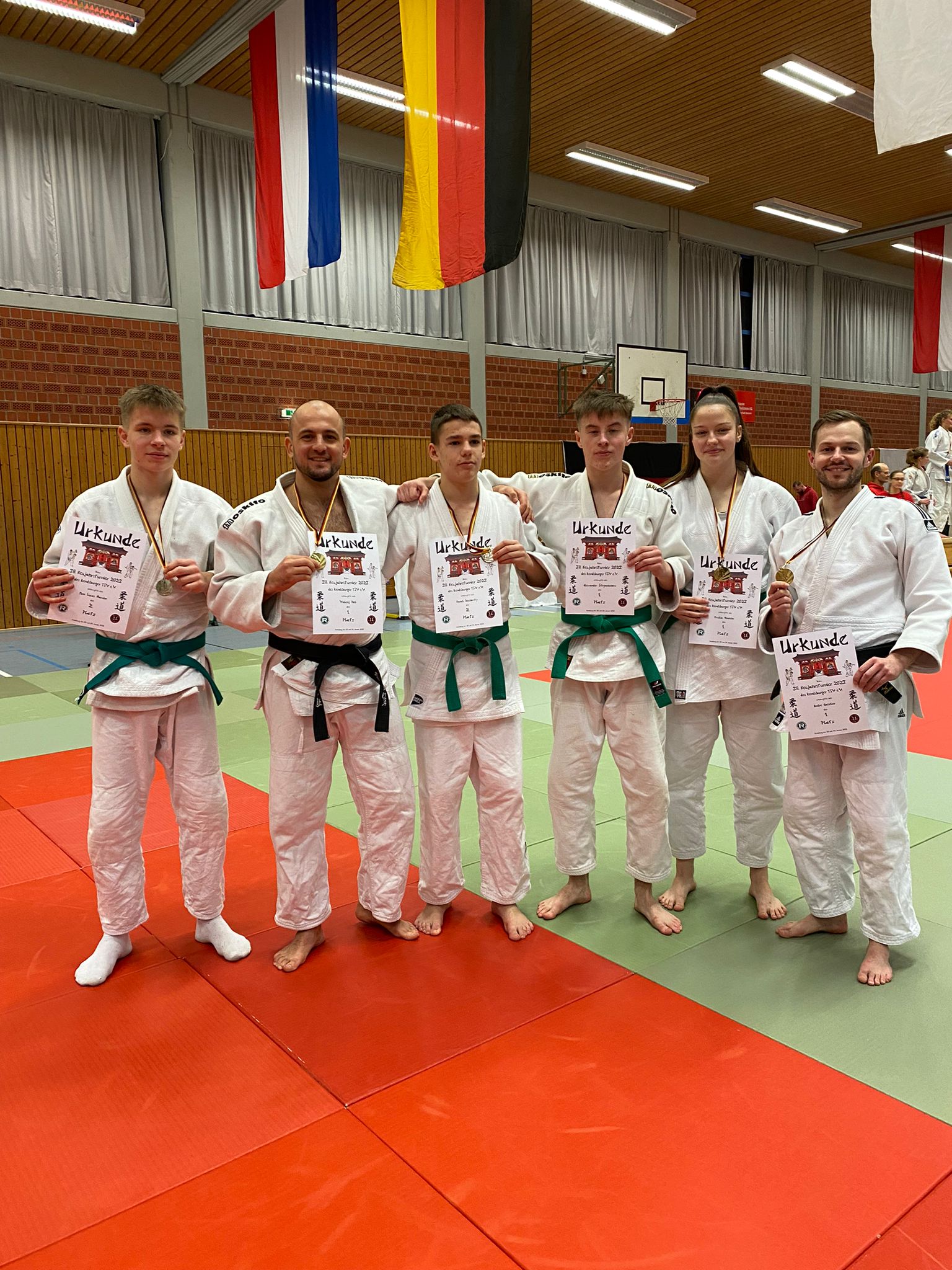 Sechsergruppe in Judokleidung hält Urkunden; Hintergrund mit Landesflaggen in einer Sporthalle.