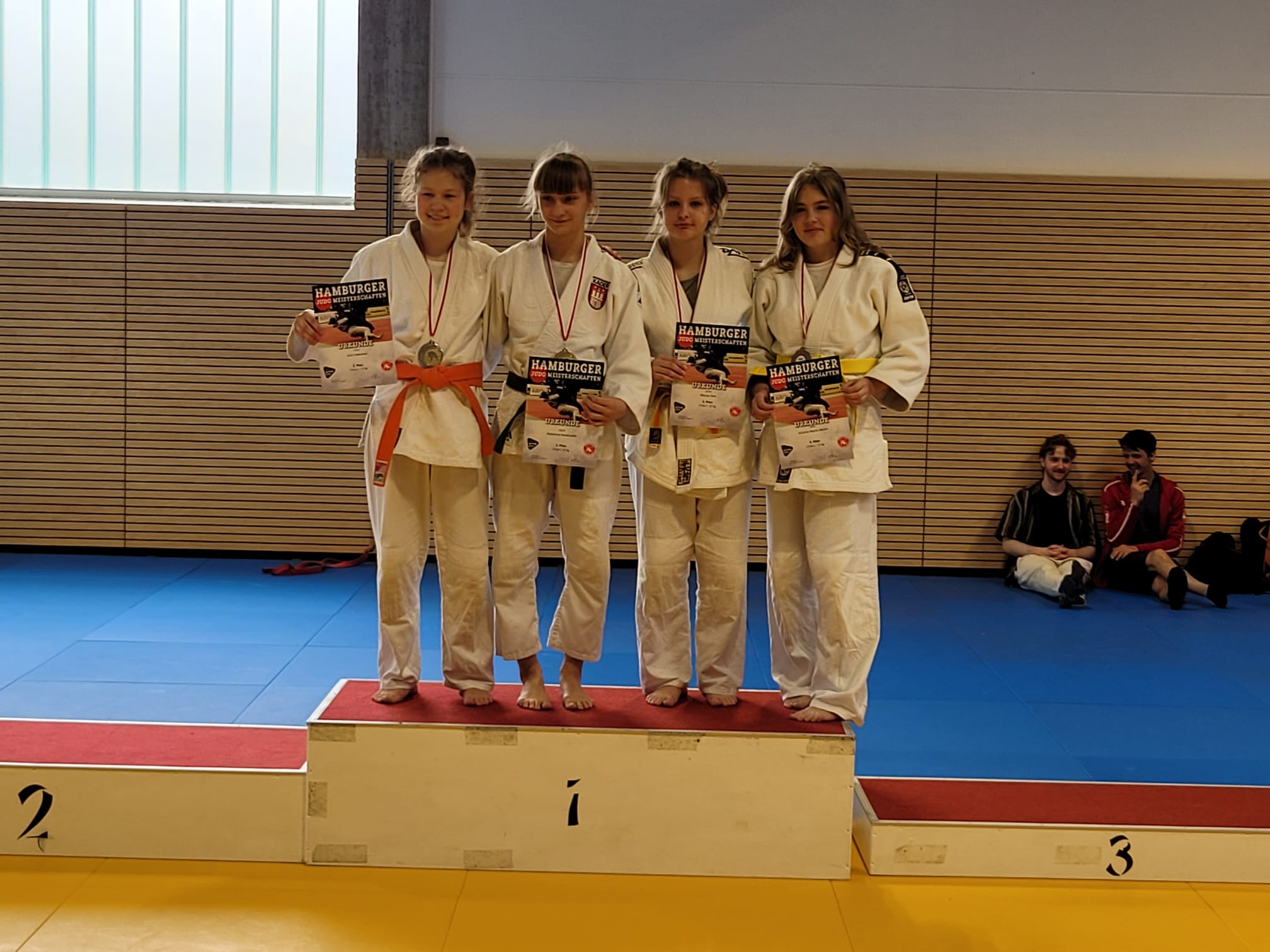 Vier Judoka auf dem Siegertreppchen halten Medaillen und Urkunden nach einem Wettkampf in einer Turnhalle.