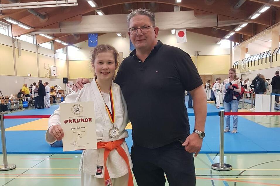 Junge in Karateanzug mit Medaille und Urkunde steht neben einem Mann in Sportbekleidung in einer Sporthalle.