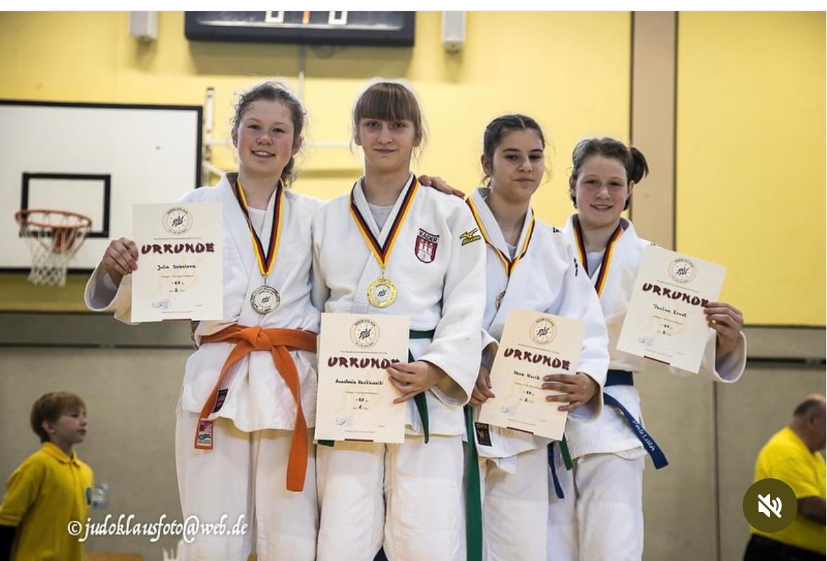 Vier Judoka in weißen Anzügen halten Urkunden und Medaillen, stehen vor einer Sporthalle mit Basketballkorb im Hintergrund.