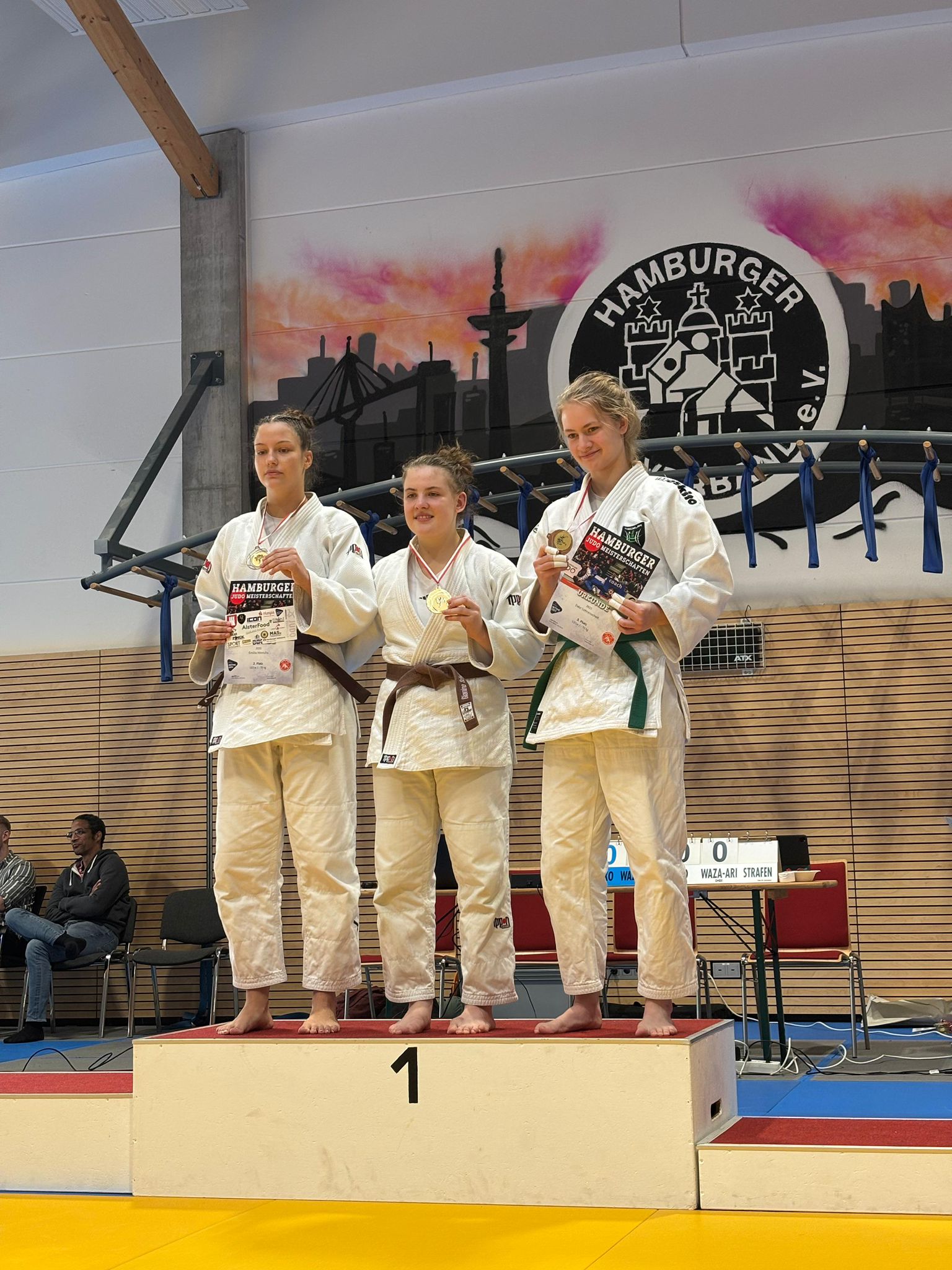 Drei Judokas auf dem Siegertreppchen, jede hält Medaillen und Urkunden in einer Sporthalle mit Hamburger Wandmalerei.