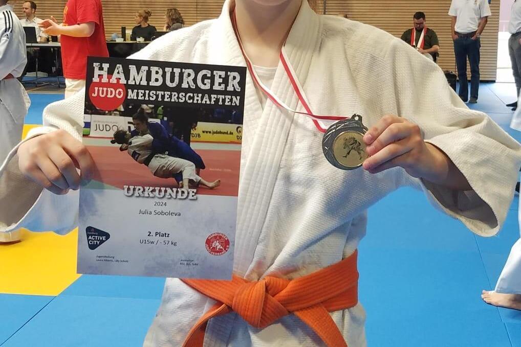 Lächelndes Mädchen in Judogi mit orangefarbenem Gürtel, hält eine Urkunde und Medaille der Hamburger Judo Meisterschaften.