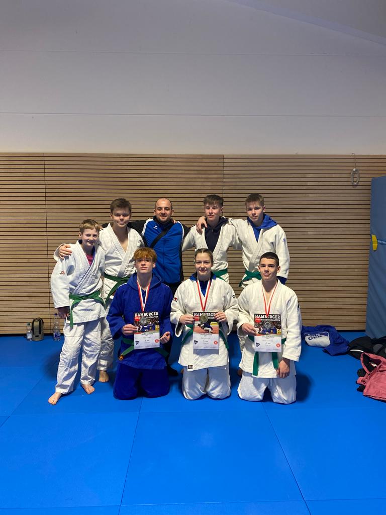 Gruppe junger Judoka in Traininganzügen mit Medaillen und Urkunden auf einer blauen Matte in einem Sportraum.