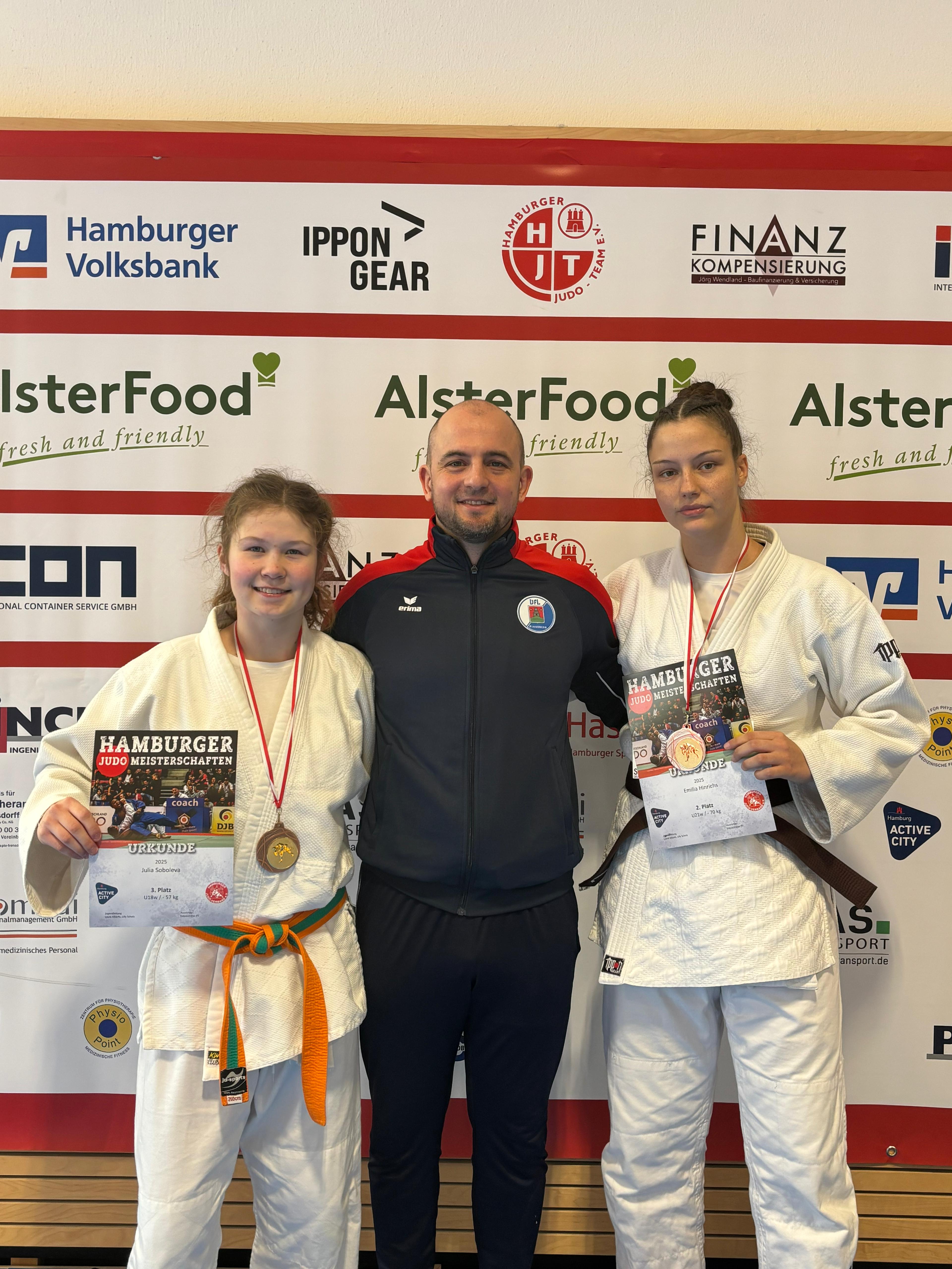 Zwei judosportliche Medaillengewinnerinnen mit ihren Urkunden stehen neben einem Trainer vor einem Werbebanner.
