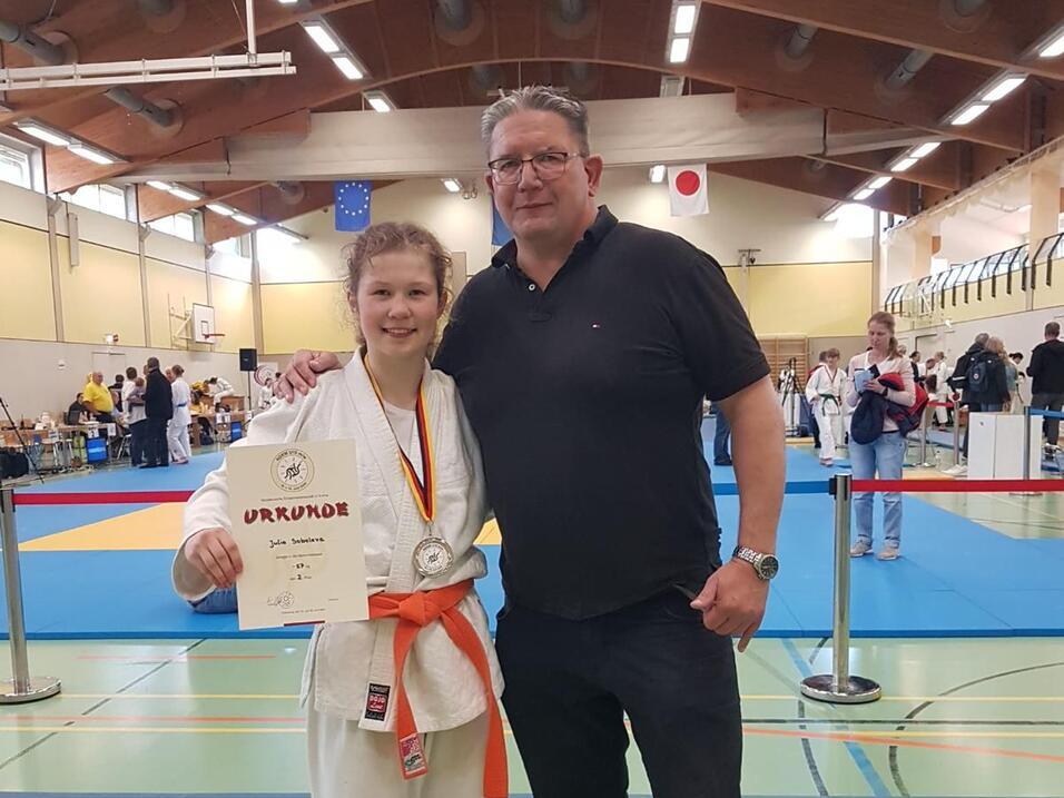 Junge in Karateanzug mit Medaille und Urkunde steht neben einem Mann in Sportbekleidung in einer Sporthalle.