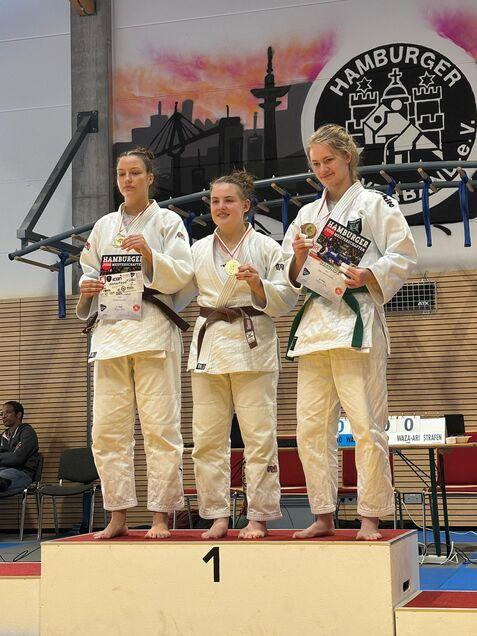 Drei Judokas auf dem Siegertreppchen, jede hält Medaillen und Urkunden in einer Sporthalle mit Hamburger Wandmalerei.