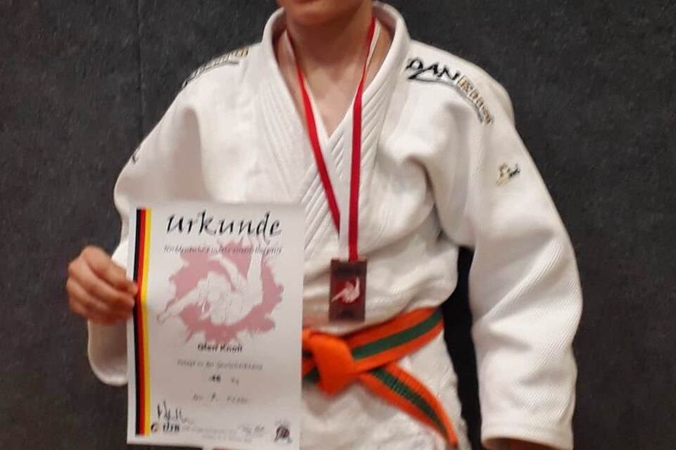 Ein junger Judoka in wei&szlig;em Gi h&auml;lt stolz eine Urkunde und eine Medaille vor einem grauen Hintergrund.