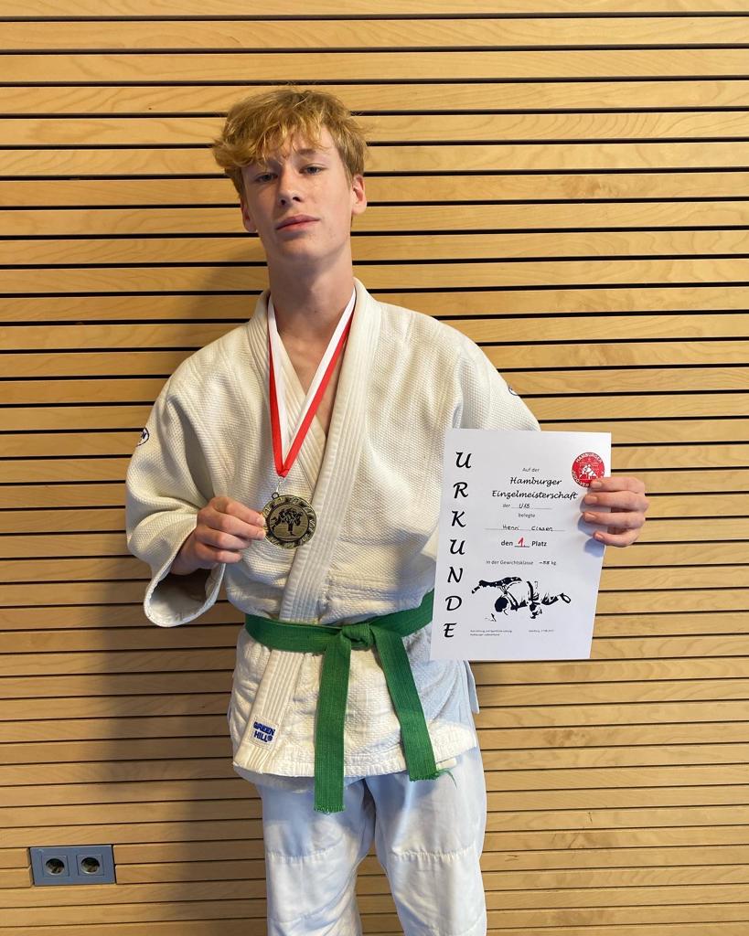 Junger Judoka in weißem Gi mit grüner Gürtelschnalle, zeigt Medaille und Urkunde für den ersten Platz bei Meisterschaft.