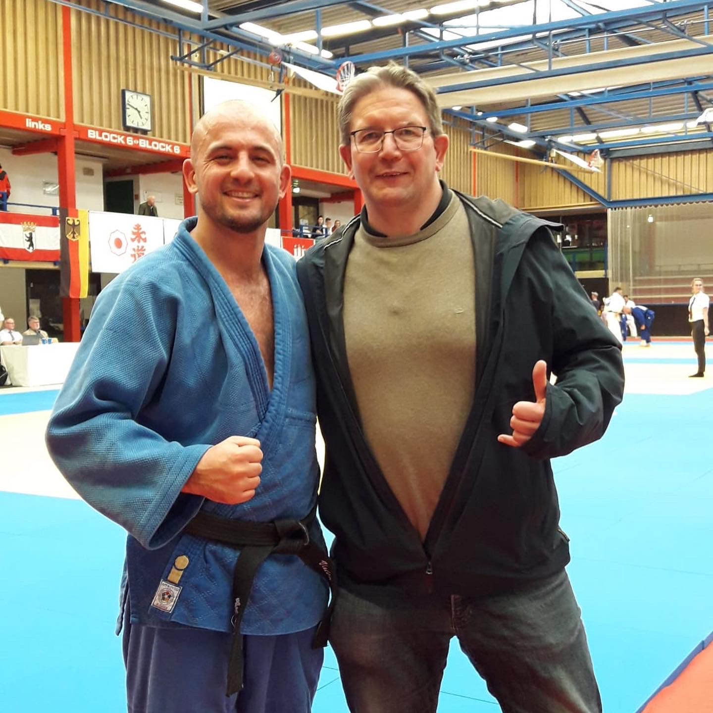 Zwei Männer posieren in einer Sporthalle, einer trägt einen blauen Judogi mit schwarzem Gürtel, der andere eine graue Jacke.