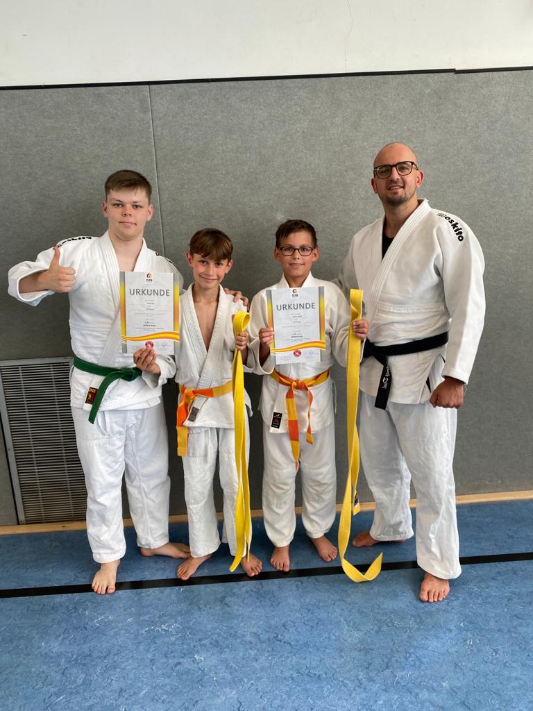 Vier Personen in Judo-Anzügen halten Urkunden und gelbe Gürtel, stehen vor einer grauen Wand und lächeln.