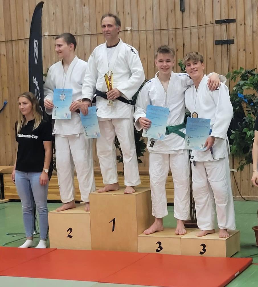 Vier Sportler in Judogi stehen auf einem Siegertreppchen, zwei halten Urkunden und der erste Platz hat einen Pokal.