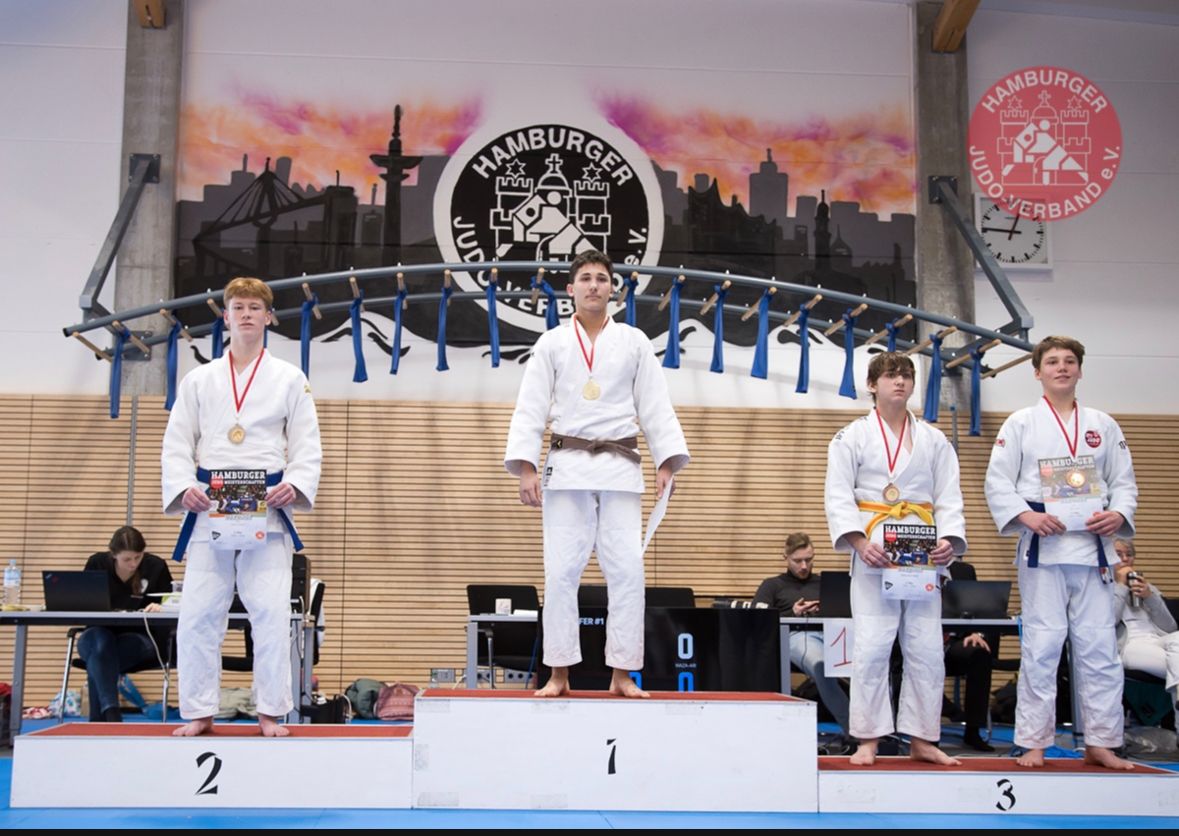 Drei Judoka stehen auf Siegertreppen, der Mittelste mit Goldmedaille, im Hintergrund das Hamburger Wappen und eine Zuschauerbank.