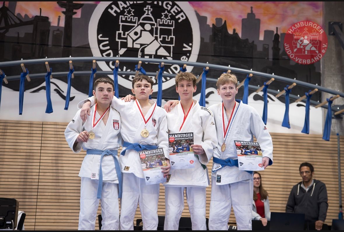 Vier junge Judokas in weißen Gis mit Medaillen stehen zusammen, halten Urkunden in einer Sporthalle mit Wandgemälde.