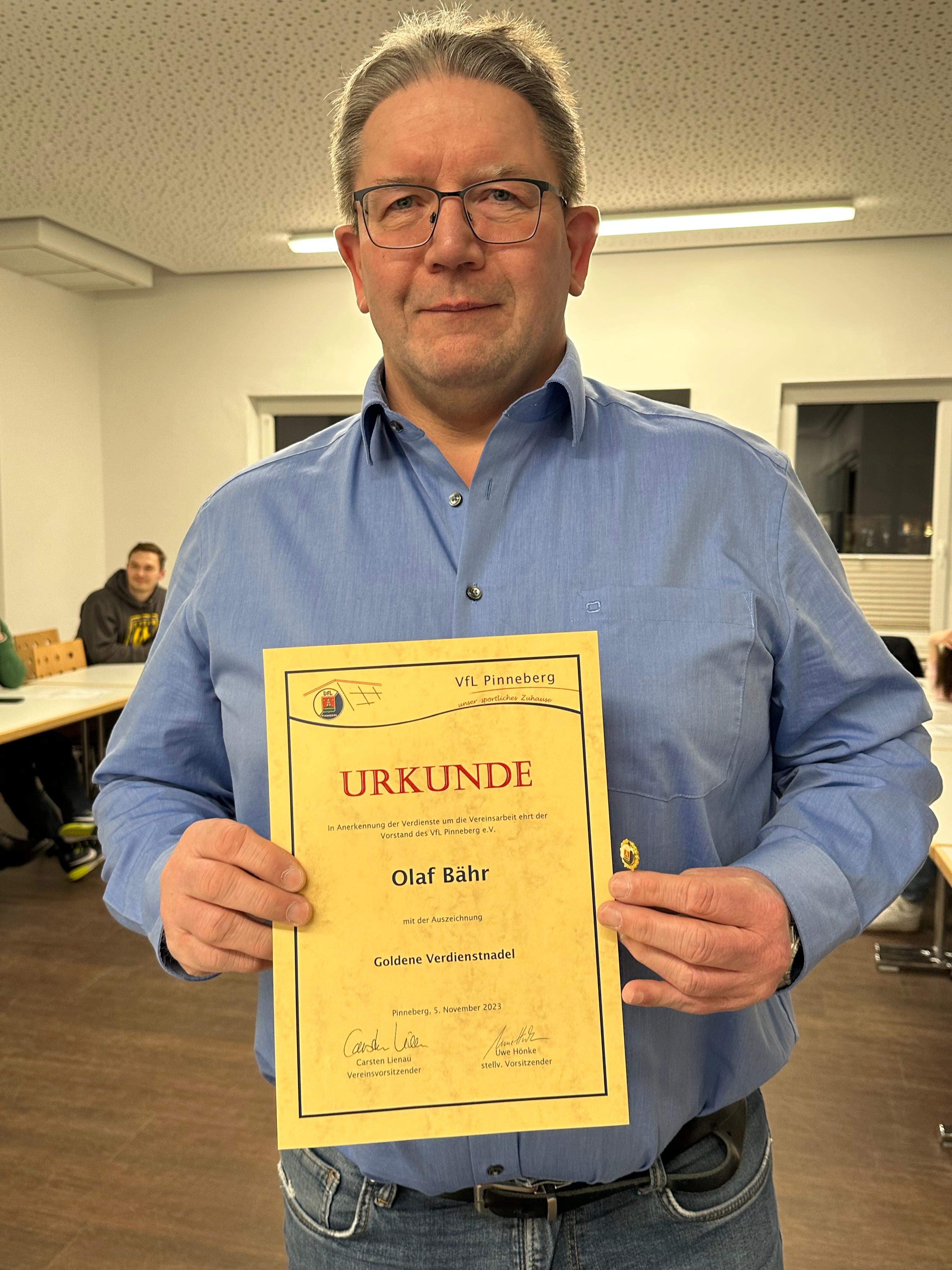 Mann in blauer Hemd hält eine Urkunde mit der Aufschrift „Goldene Verdienstnadel“ in einem Vereinsraum des VfL Pinneberg.