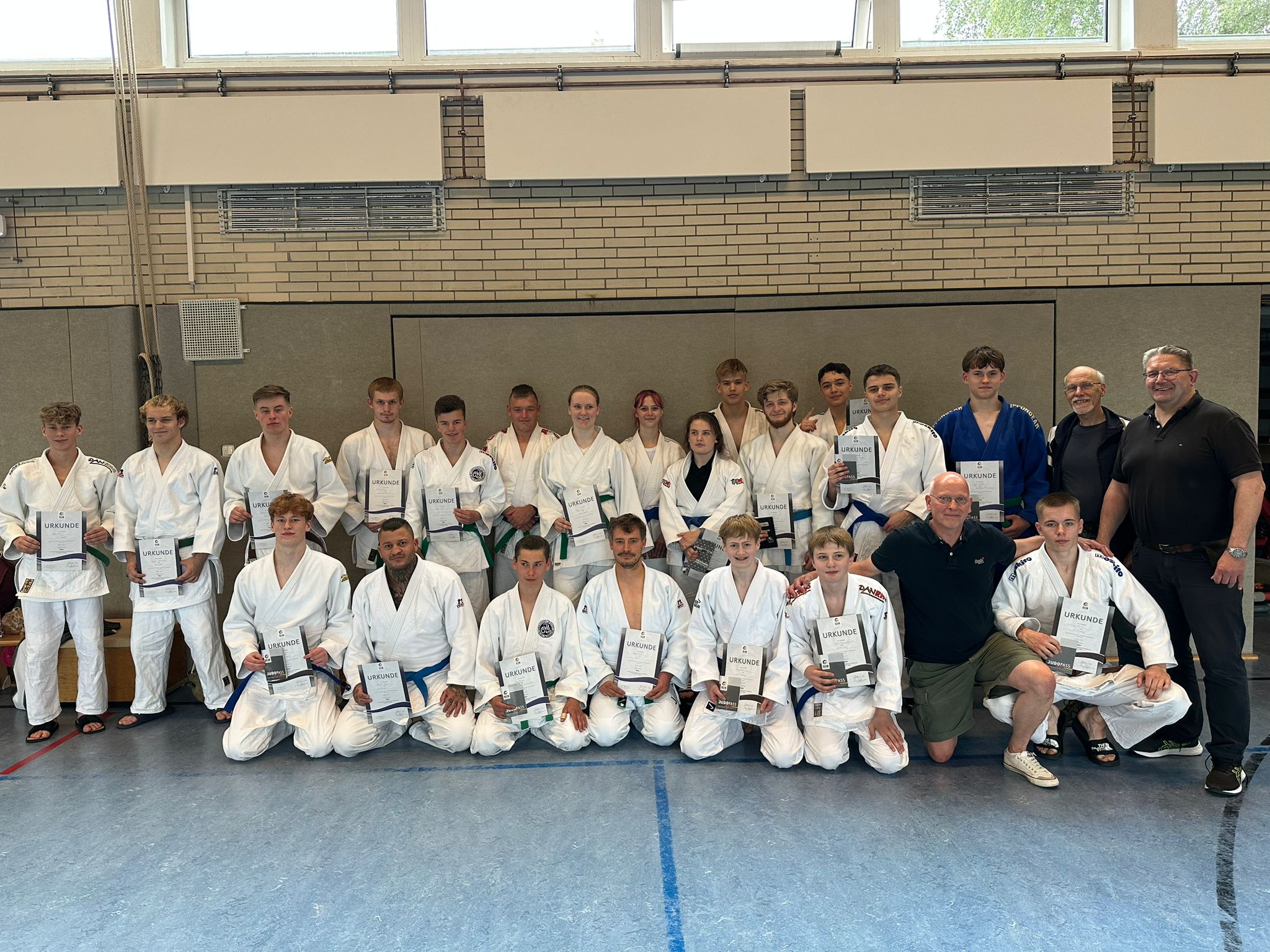 Gruppe von Judokas in weißen Anzügen, die stolz ihre Urkunden in einem Sportraum halten.