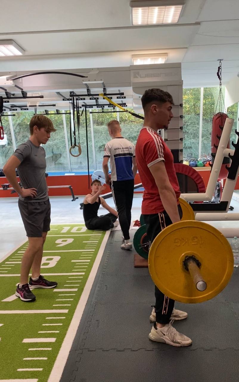 Vier Jugendliche in einem Fitnessstudio; einer hebt eine Langhantel, andere beobachten oder trainieren.