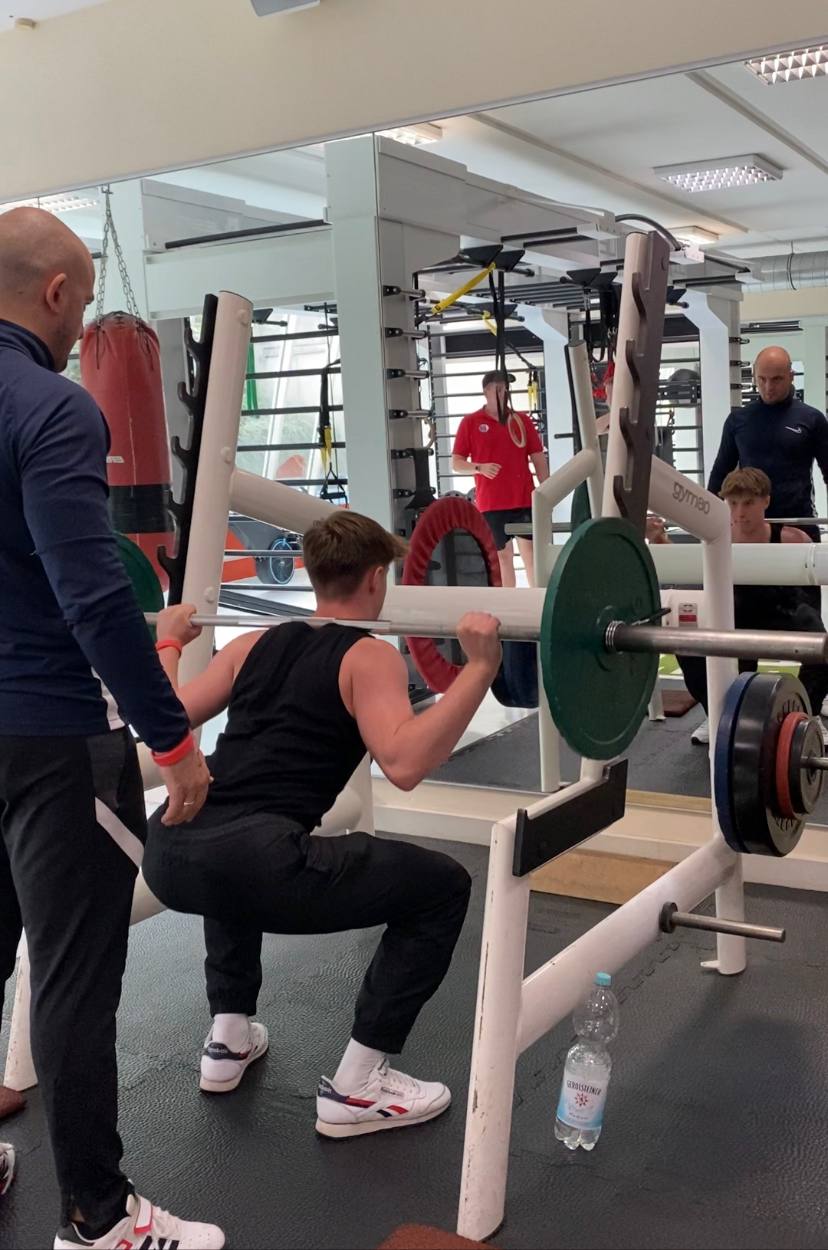 Junger Mann beugt sich mit einer Langhantel auf der Schulter in einer Fitnessstudio-Umgebung.