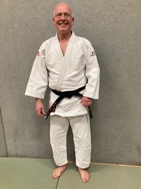 Lächelnder Mann in Judoanzug mit schwarzem Gürtel steht barfuß vor grauem Hintergrund.