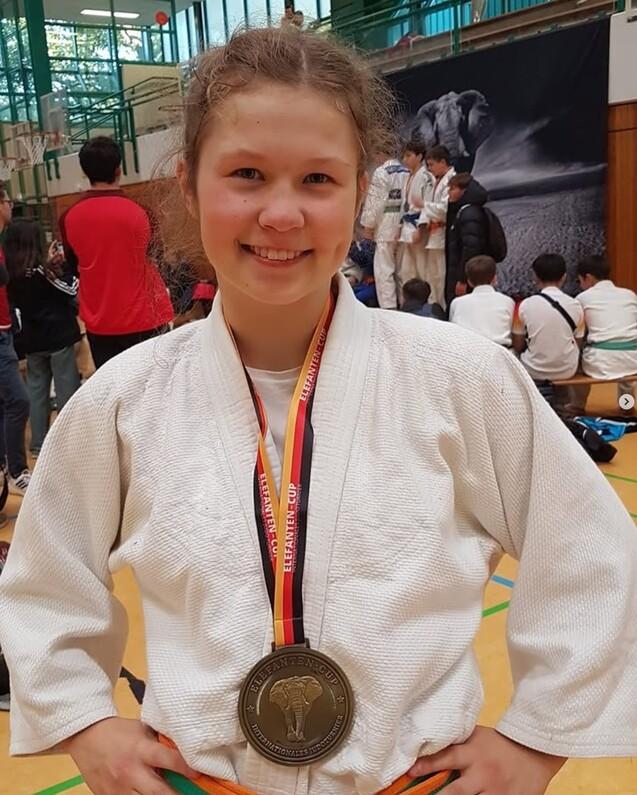 Junge Judoka in weißem Gi mit Medaille um den Hals lächelt auf einem Wettkampf im Hintergrund.