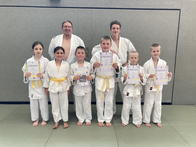 Gruppe von Kindern in Judogis mit Urkunden und einem Trainer in einem Trainingsraum.