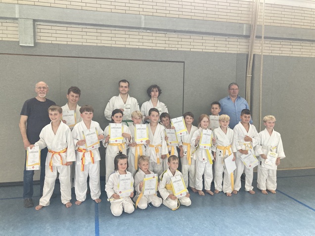 Gruppe von Kindern und Erwachsenen in weißen Karateanzügen mit Urkunden in einer Turnhalle.