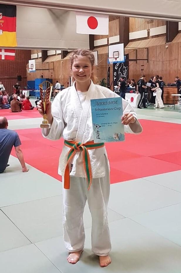 Junge im Judogi hält einen Pokal und eine Urkunde für den ersten Platz auf einem Turnier, im Hintergrund Judokämpfe.