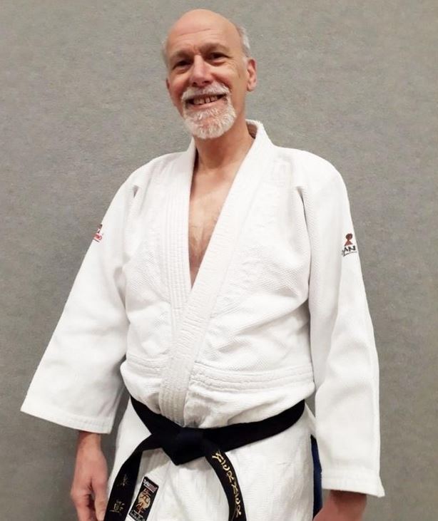 Ein lächelnder Mann in einem weißen Judoanzug mit schwarzem Gürtel steht vor einer grauen Wand.