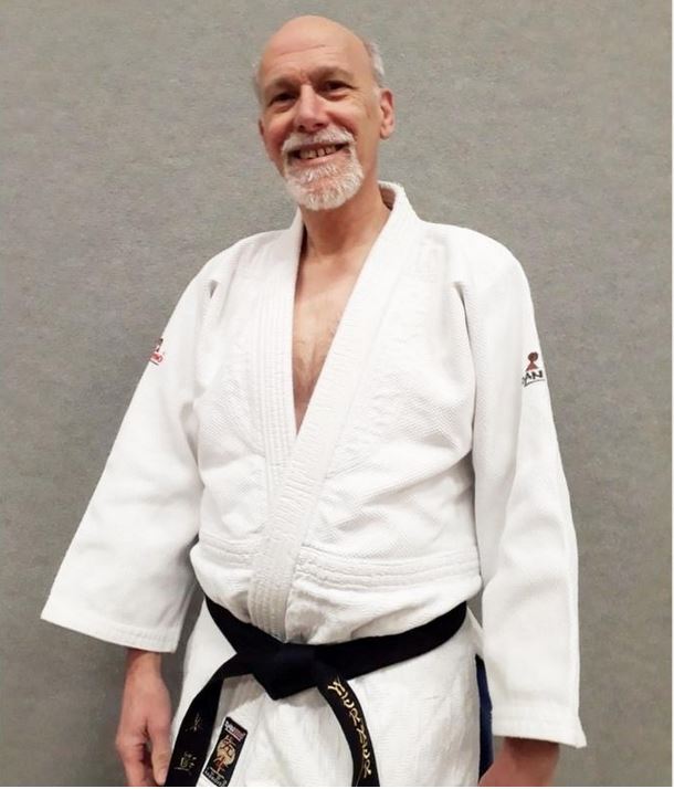 Lächelnder Mann mit grauem Bart im weißen Judoanzug und schwarzem Gürtel vor grauem Hintergrund.