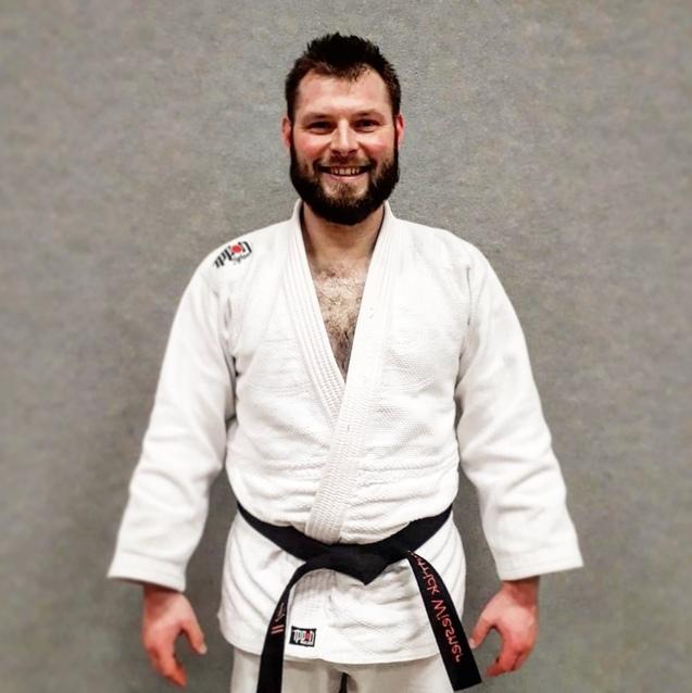 Lächelnder Mann in weißem Judogi mit schwarzem Gürtel vor grauem Hintergrund.