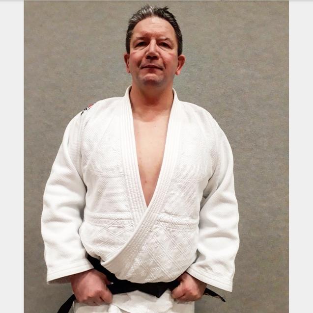 Mann in einem weißen Judo-Gi mit schwarzem Gürtel steht vor grauem Hintergrund und blickt direkt in die Kamera.